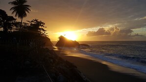 A beach scene with a sunset, palm trees, and rocky coastline. - Birdie's Nest condo #2 with ocean view (Black Rock)