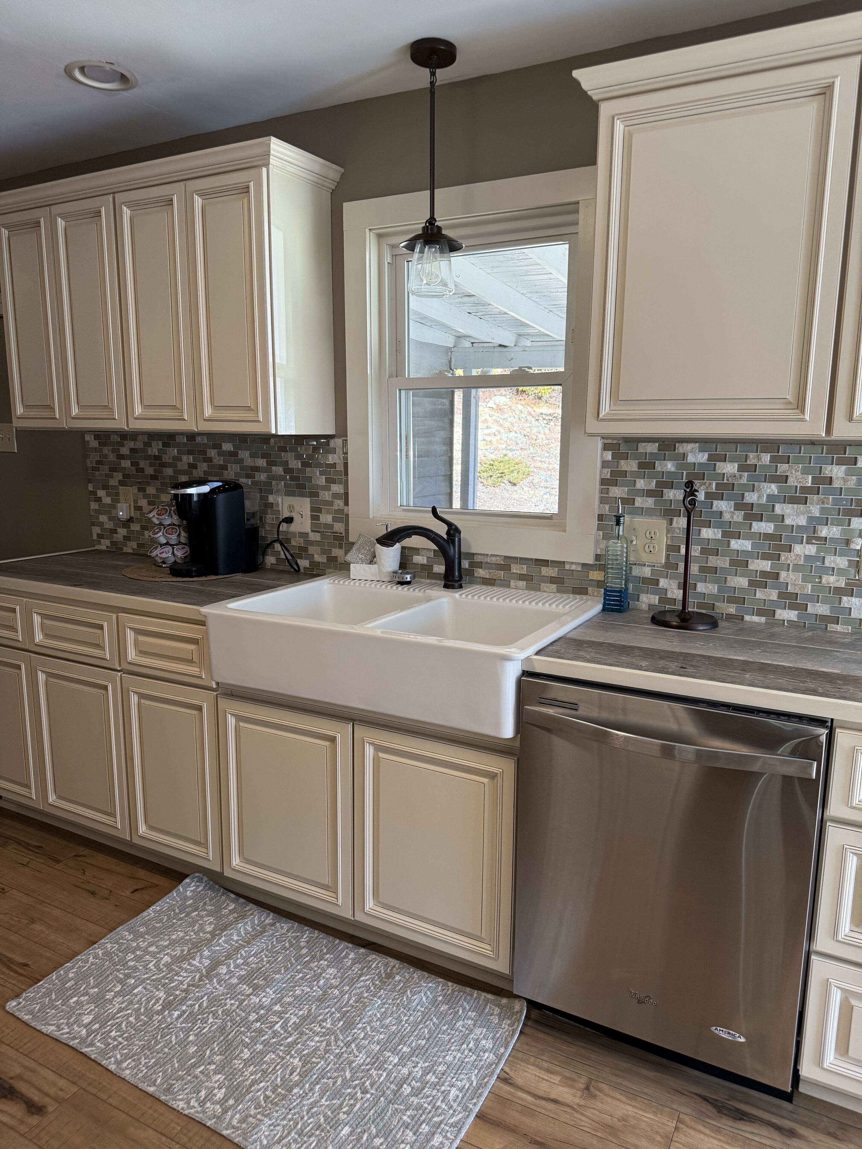 A kitchen with beige cabinetry, a stainless steel dishwasher, a white sink, and a window with a view of the outdoors.