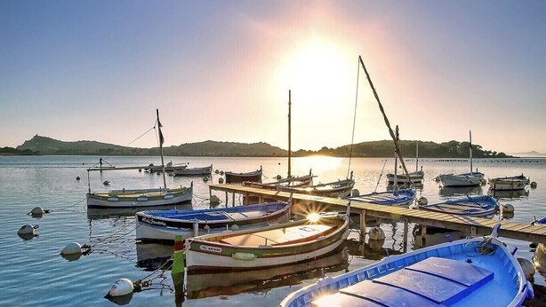 Un port de plaisance où plusieurs bateaux sont amarrés, un ciel dégagé et un soleil se couchant à l’horizon.