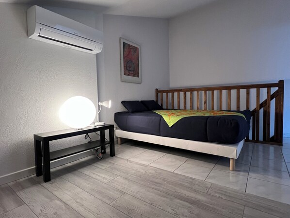 Een slaapkamer met een bed, nachtkastje met een lamp, een airconditioner en een ingelijst schilderij aan de muur.