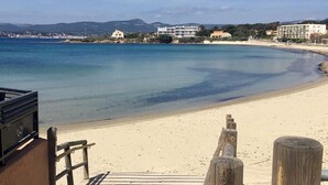 Une promenade en bois mène à une plage de sable fin, bordée d’une eau d’un bleu limpide, avec des bâtiments visibles au loin.