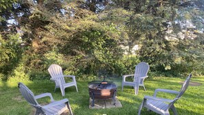 Un foyer est entouré de quatre chaises Adirondack dans un espace extérieur verdoyant, avec des arbres en arrière-plan.