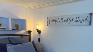 Une chambre à coucher avec un lit, une table de chevet avec une lampe, une étagère avec des livres et une plante, et un panneau mural portant les mots « reconnaissant(e), plein(e) de gratitude, béni(e) ».