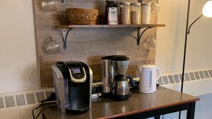Un coin cuisine équipé d’une machine à café, d’une bouilloire et de bocaux de rangement placés sur une étagère en bois.