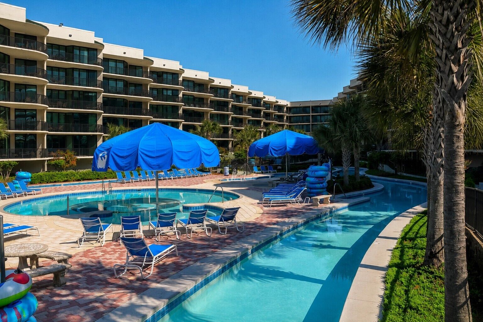 Una struttura alberghiera con area piscina, diversi ombrelloni blu e poltrone relax.