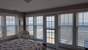 Un dormitorio con una cama grande, una silla de mimbre y vistas al océano a través de ventanas amplias con persianas.