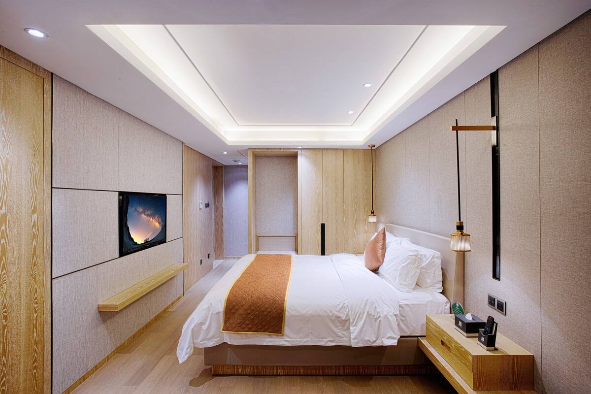 A modern hotel room with a large bed, a TV mounted on the wall, a bedside table with a lamp, and a wooden bench.