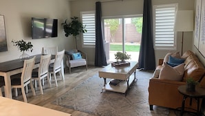 A modern living room with a dining area, a large window with blinds, a ceiling fan, and a flat-screen TV mounted on the wall. - Pool Side Villa |Pet-Friendly Modern Escape w/ Games, Pool Toys, Bikes & More! Near St.George (Washington)