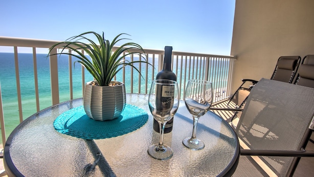 A round table with a potted plant, a wine bottle, and two wine glasses on a balcony overlooking the ocean.