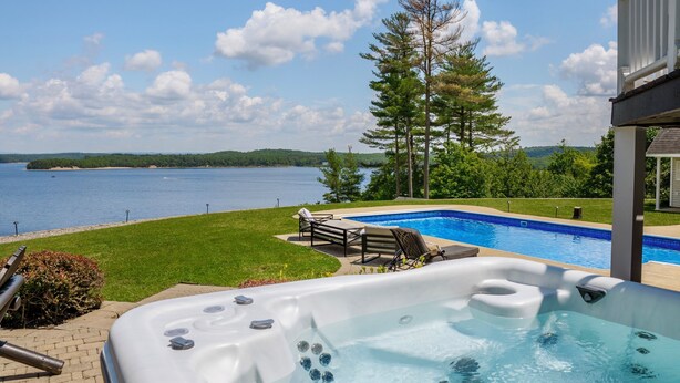 A hot tub with a view of a lake, surrounded by a deck and a lounge chair.