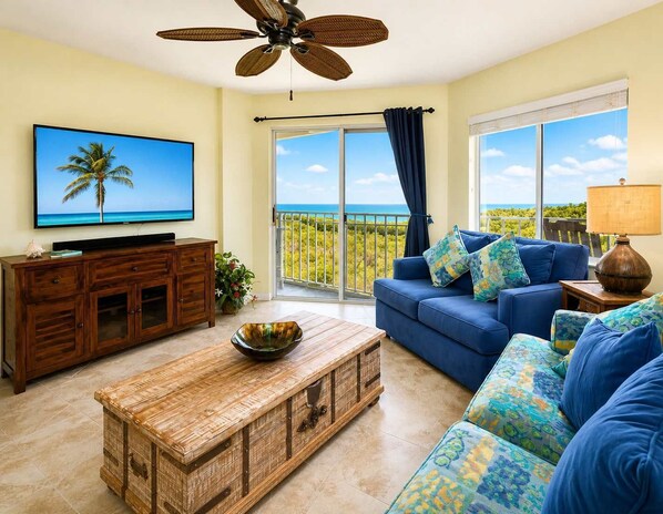 A living room with a television, a sofa, a coffee table, and a view of the ocean through large windows.