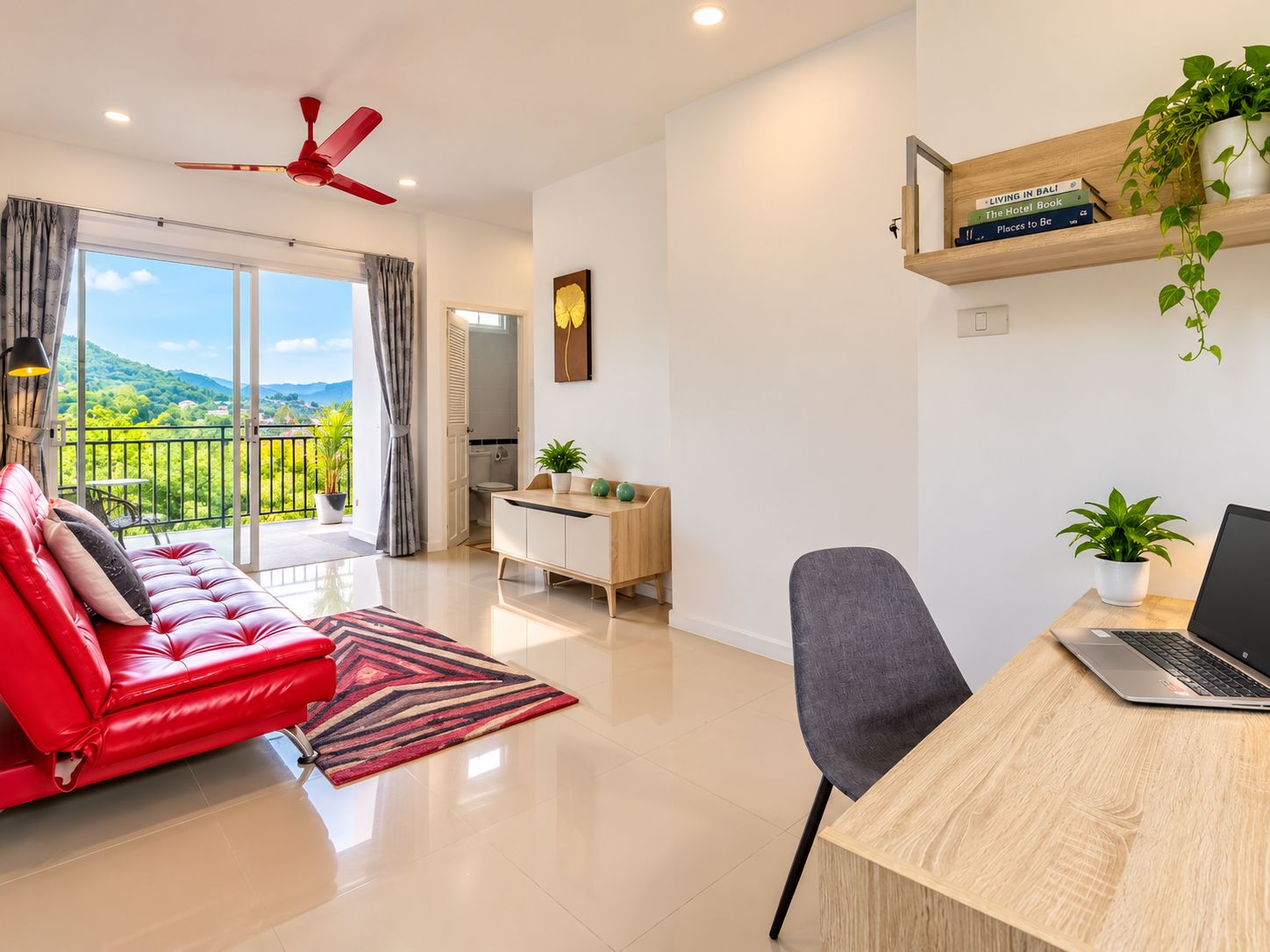 A modern living room with a red sofa, a laptop on a wooden desk, and a balcony with a view of greenery.