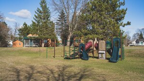 Ein Spielplatz mit verschiedenen Spielgeräten, umgeben von Bäumen und Häusern auf einer grünen Fläche.