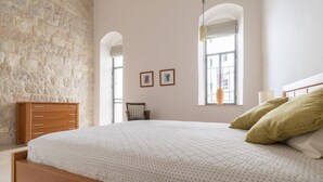 Une chambre avec un grand lit, une commode en bois, un mur en pierre et deux fenêtres avec des rideaux.