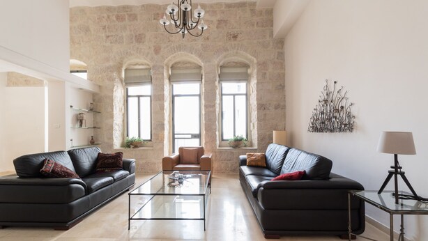 Un vaste salon agrémenté de deux canapés en cuir noir, d’une table basse en verre et d’un lustre. Il y a trois fenêtres à arc en plein cintre, et une pièce décorative en métal est fixée au mur.