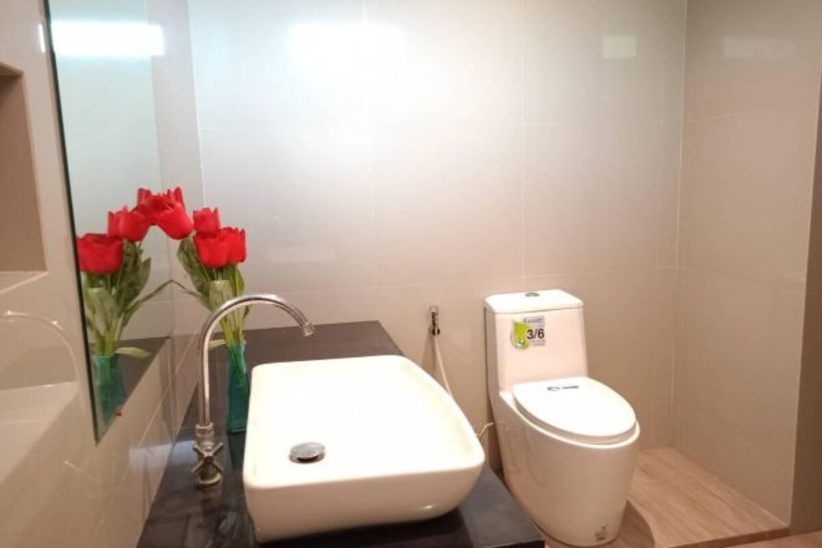 Een badkamer met een wit wastafel, een moderne kraan en een toilet. Aan de linkerkant staan rode bloemen in een vaas.