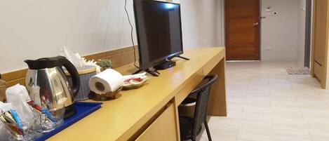 Quarto de hotel com escrivaninha de madeira, televisão, chaleira e uma cadeira.
