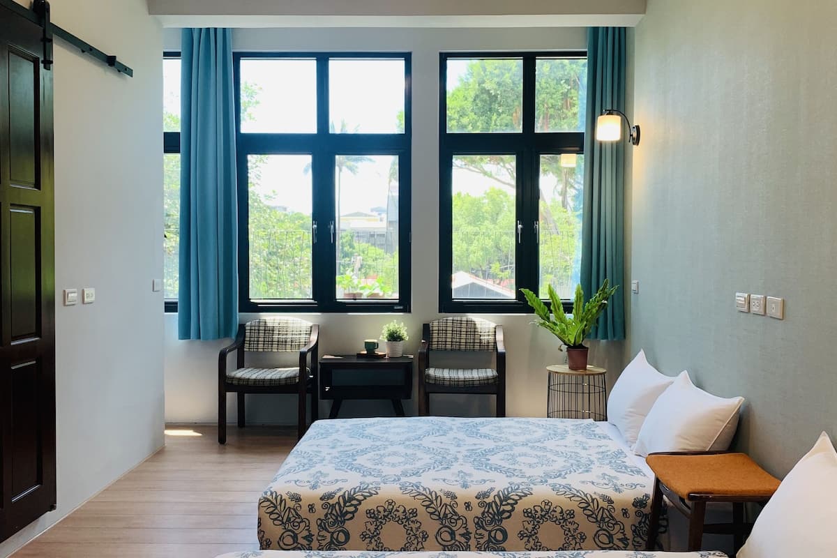 Habitación de hotel con dos camas, un escritorio, sillas, una planta en maceta y ventanas grandes con cortinas.