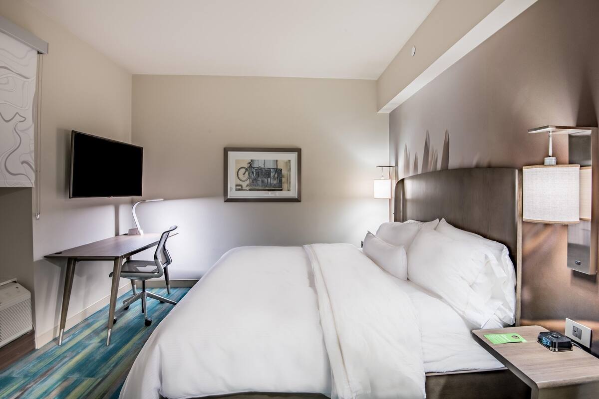 Hotellihuone, jossa on sänky, työpöytä, tuoli, televisio, taideteos ja yöpöytä, jossa on lamppu.