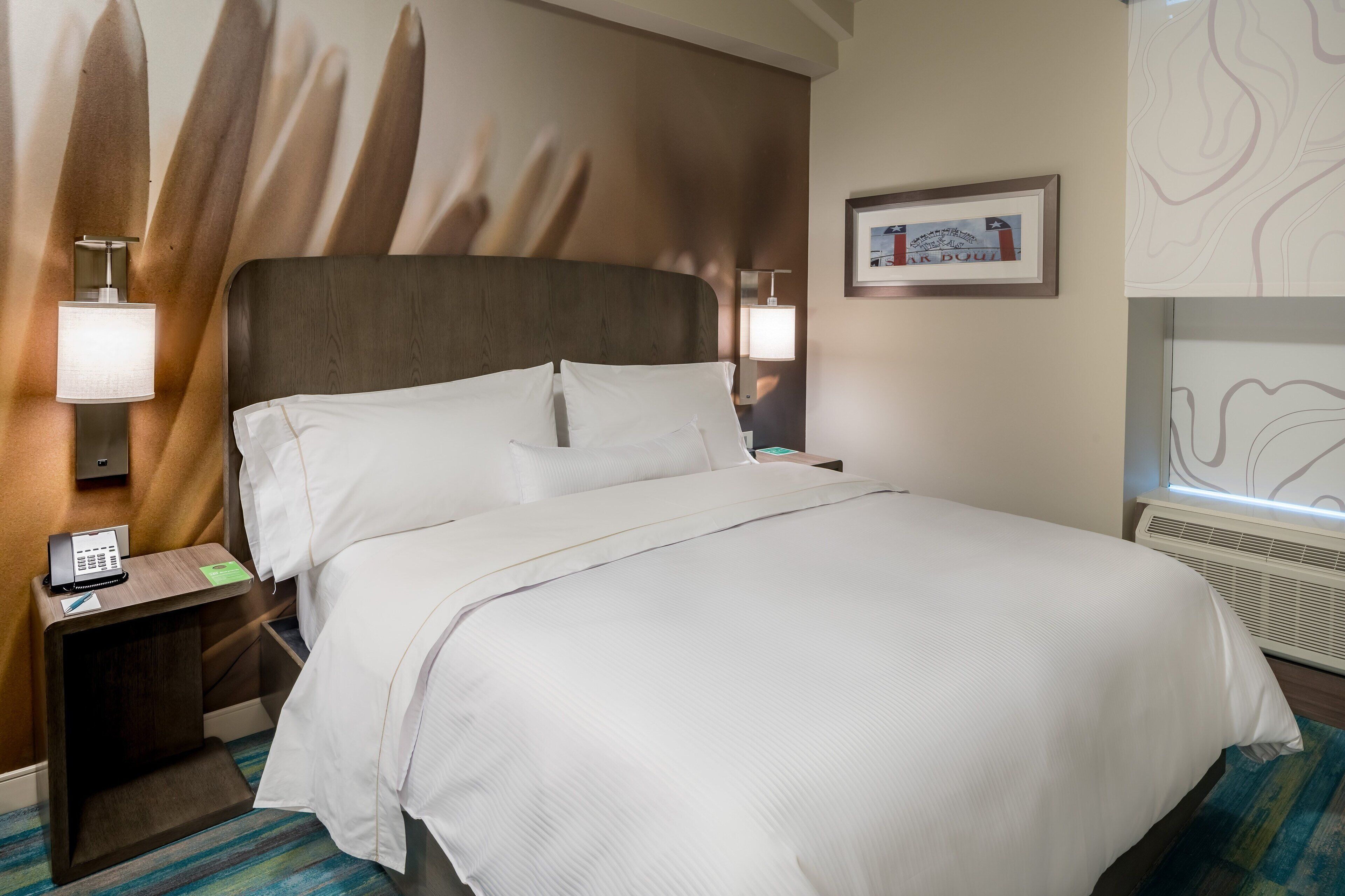 A hotel room with a large bed, a nightstand with a phone, a framed picture on the wall, and a window with a view.