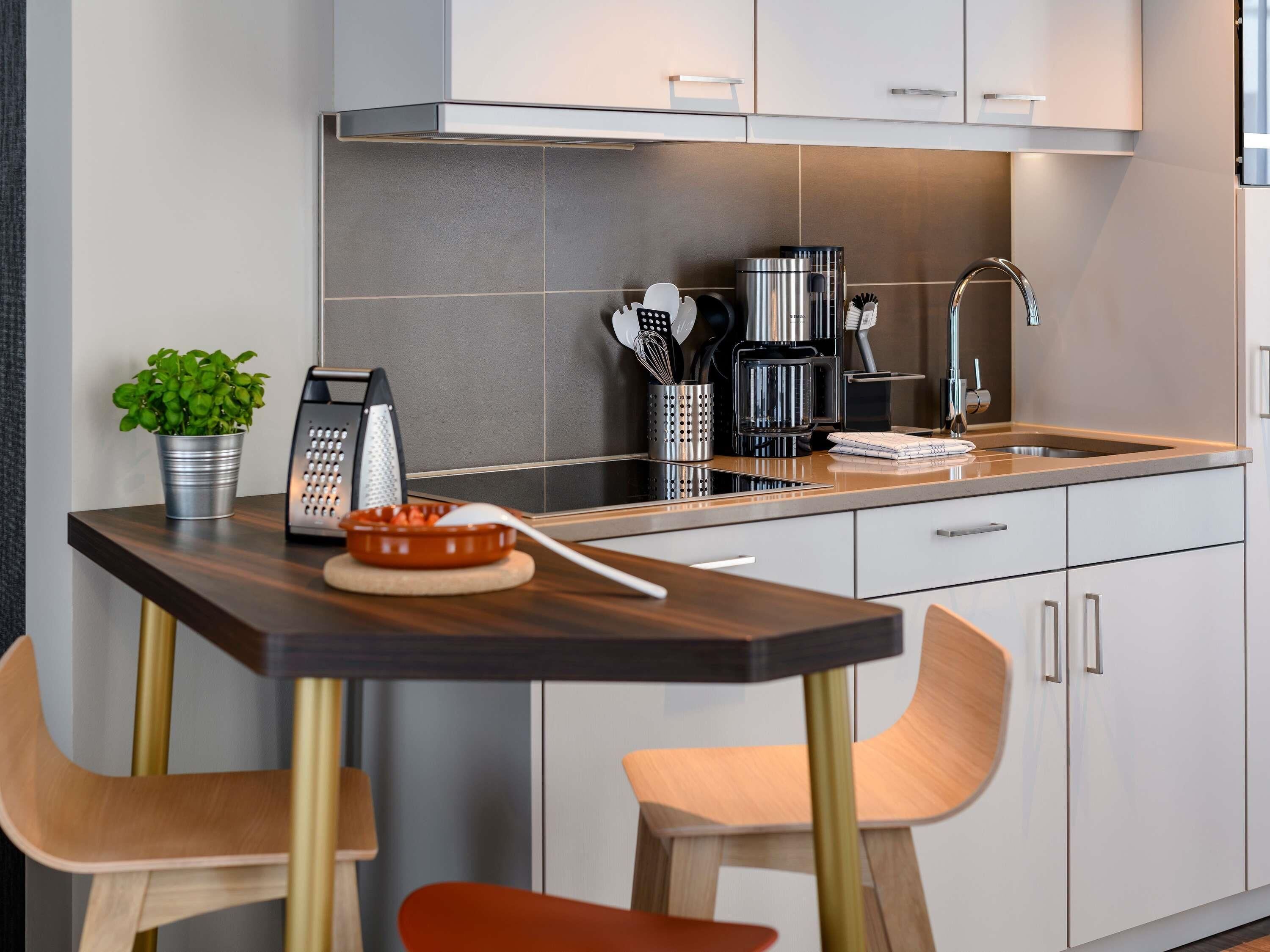 Une cuisine moderne avec un plan de travail foncé, deux chaises en bois, une bouilloire en acier inoxydable, une machine à café et une plante en pot.