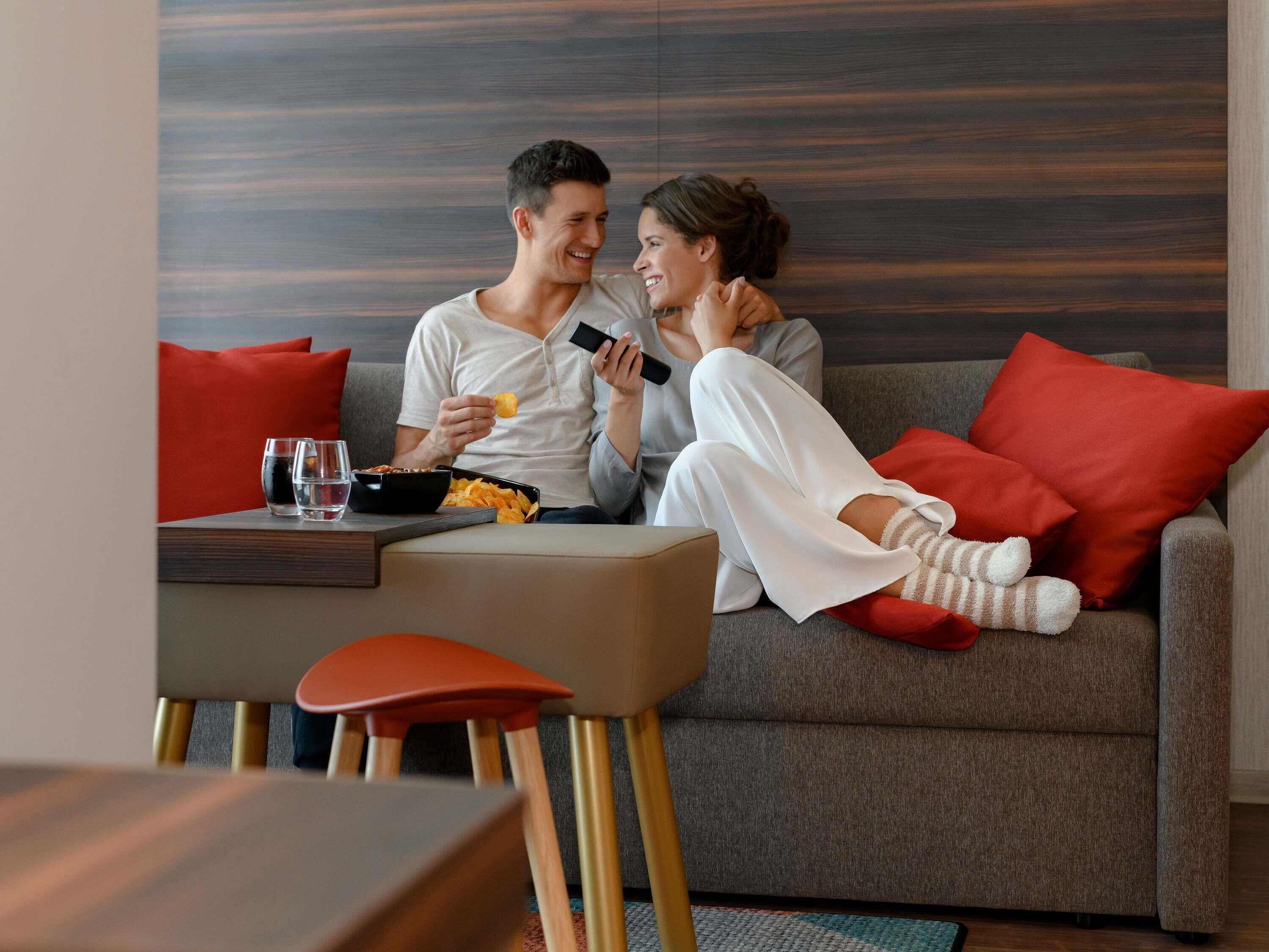 Un salon moderne avec un canapé gris, des coussins rouges, une petite table avec des verres et des en-cas, et une chaise en bois.