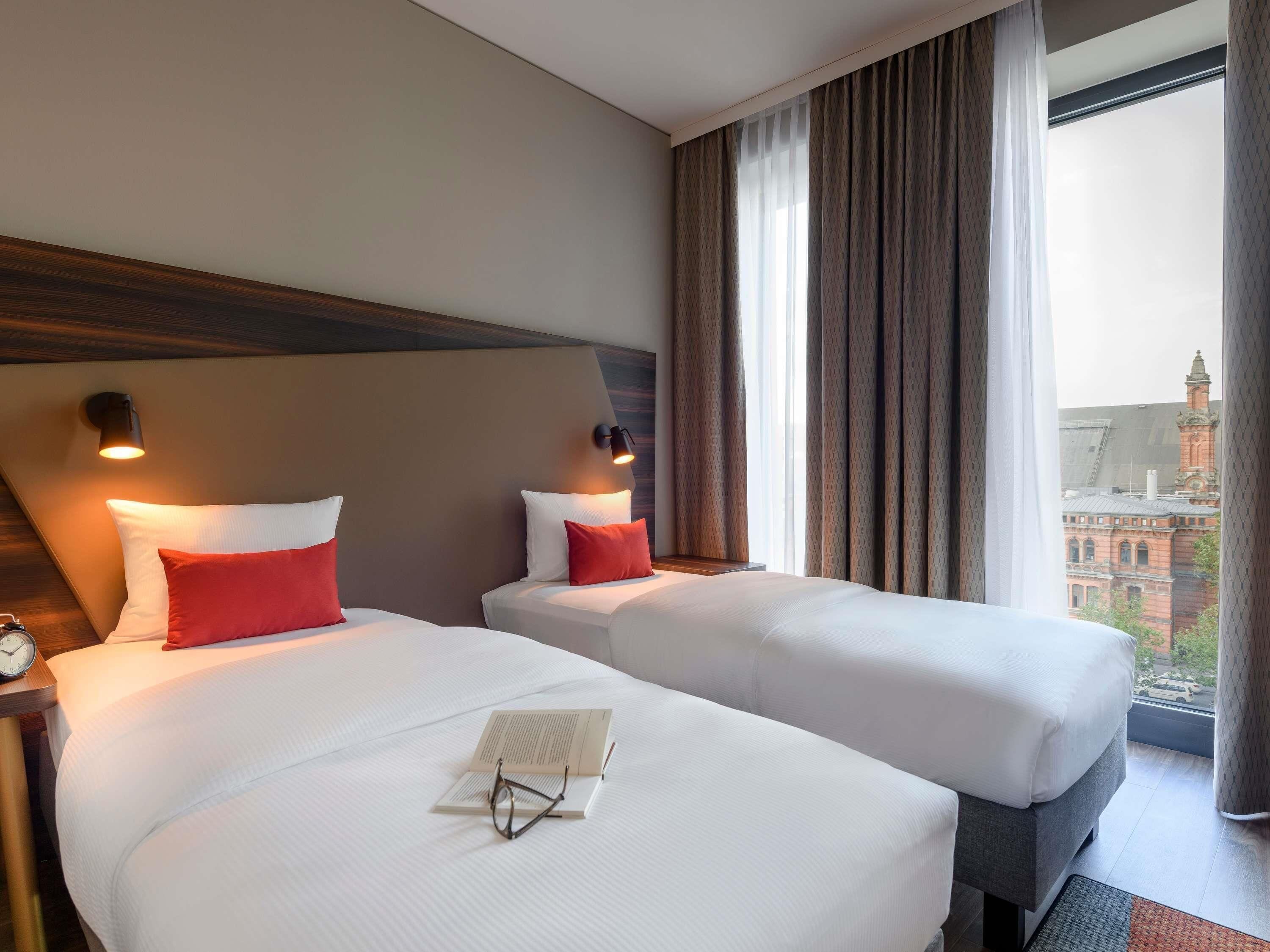 Une chambre d’hôtel avec deux lits, une table de chevet, une horloge et une vue sur un immeuble par la fenêtre.