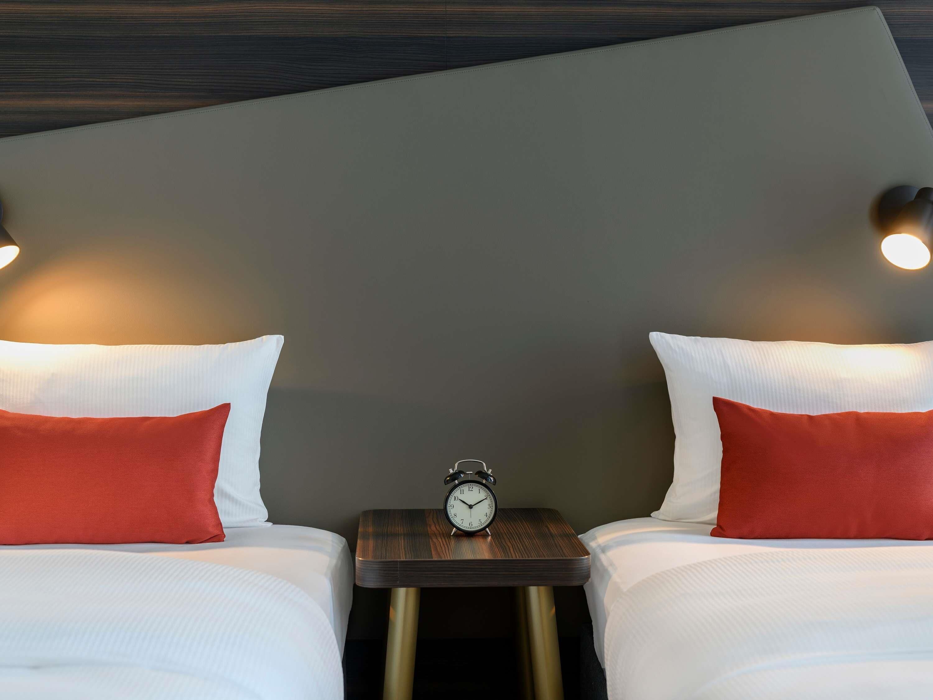 A hotel room with two beds, each with white bedding and red pillows, a small bedside table with a clock, and wall-mounted light fixtures.