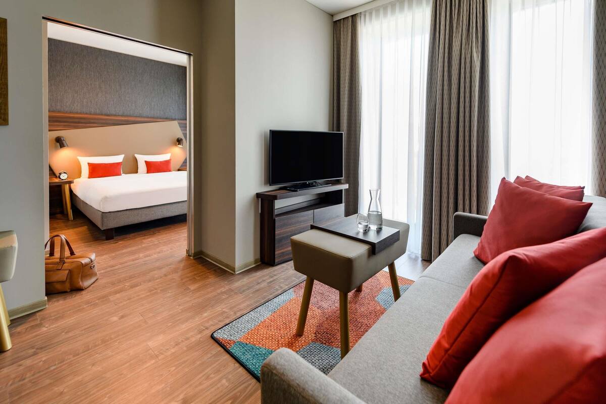 Ein Hotelzimmer mit Bett, Fernseher, Sofa, kleinem Tisch und einem Fenster mit Vorhängen.