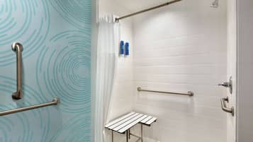 Une salle de bain avec des murs carrelés de carreaux blancs, une douche équipée d’un pommeau de douche à main et une petite étagère blanche.