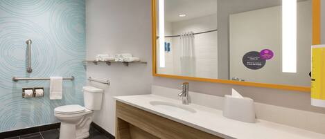 A modern bathroom with a white toilet, a wooden vanity with a sink, and a large mirror. There are handrails, a towel rack, and a shelf with toiletries.