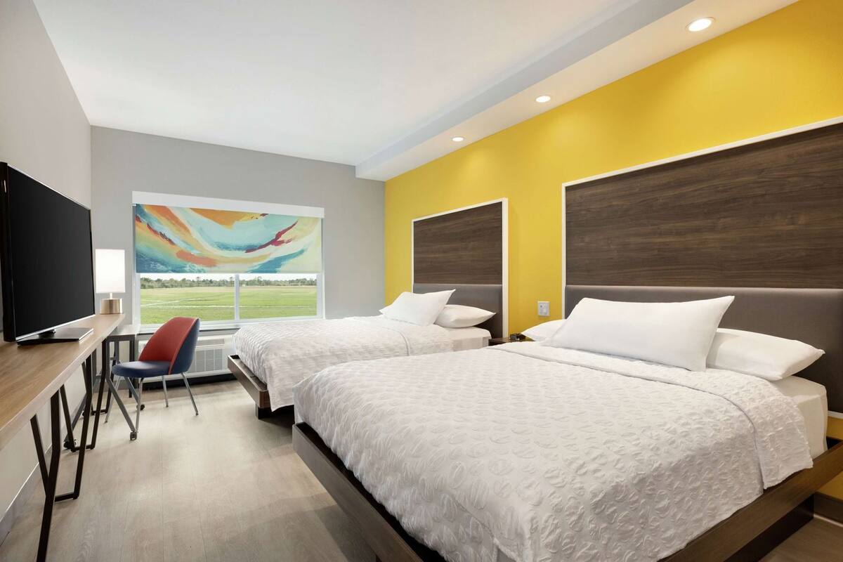 A hotel room with two beds, a desk with a chair, a television, and a large window with a view of a field.