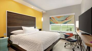 A modern bedroom with a large bed, a desk with a computer, and a large window with a view of a green field.