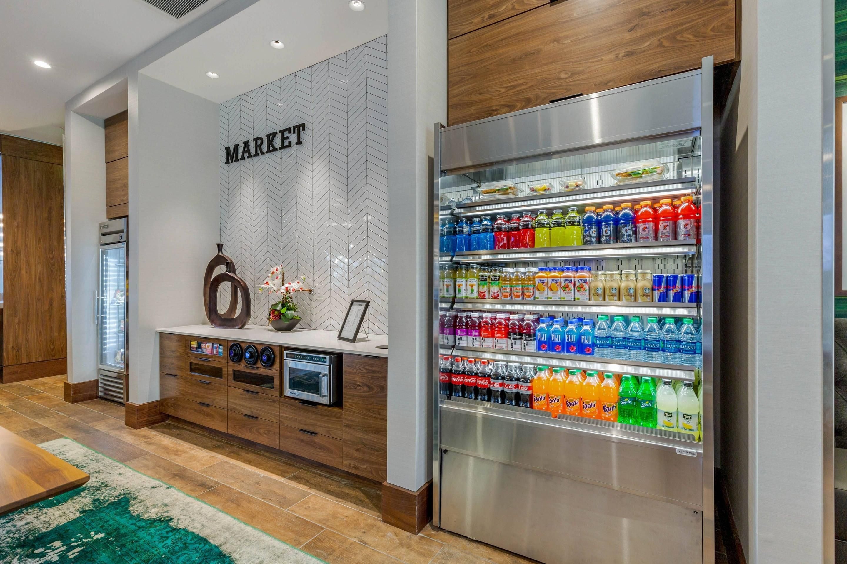 A hotel lobby with a kitchenette area, a refrigerator stocked with beverages, and a wall labeled 'MARKET'.