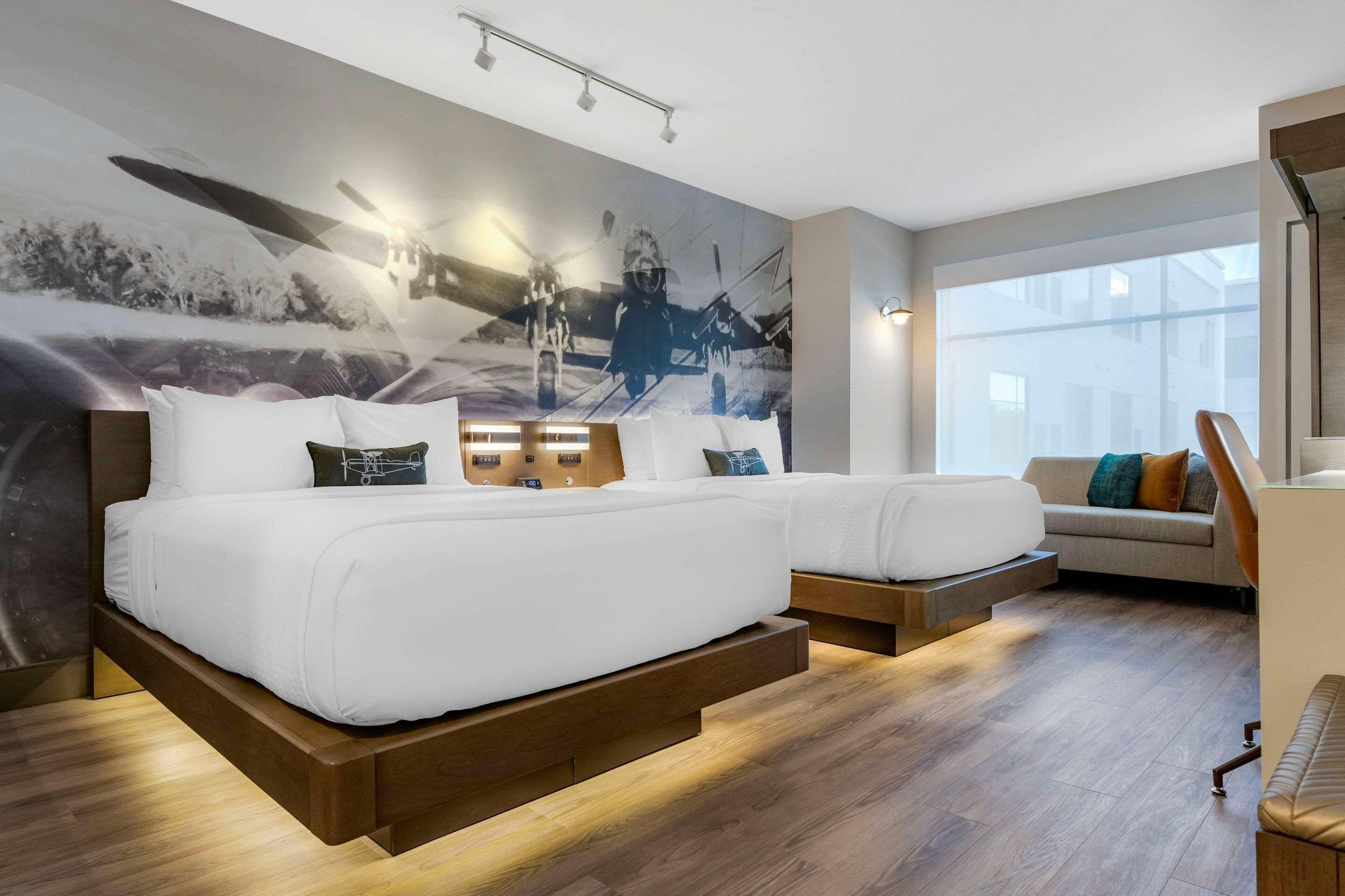 A modern hotel room with a large bed, a sofa, a wooden floor, and a large window.