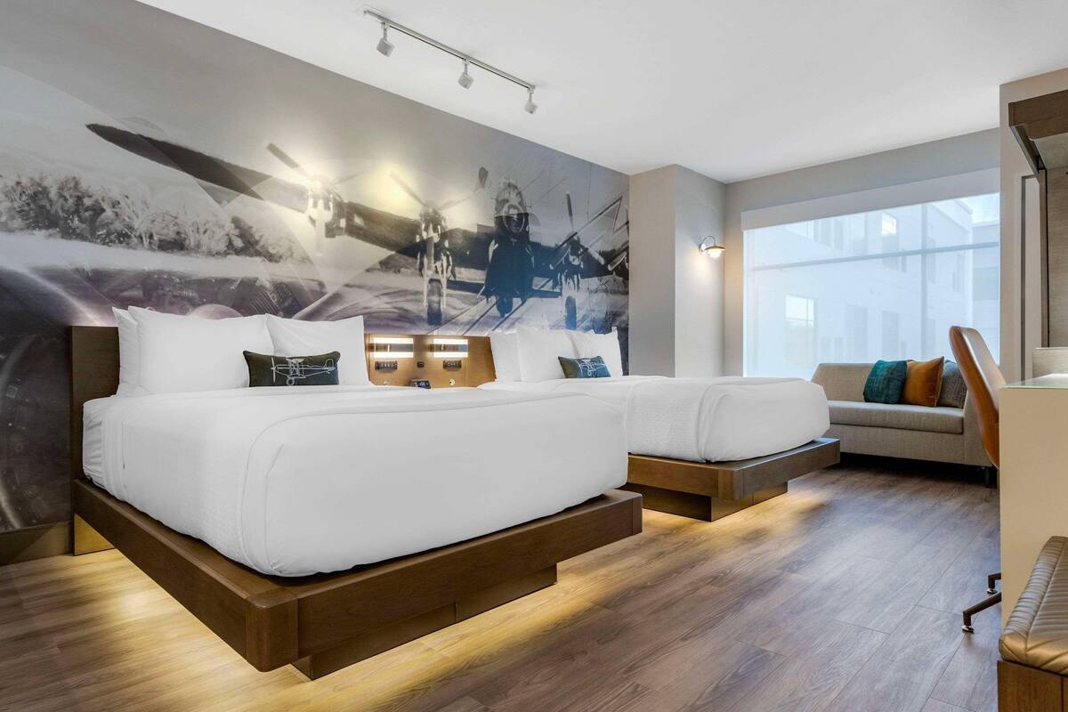 A modern hotel room with a large bed, a sofa, a wooden floor, and a large window.