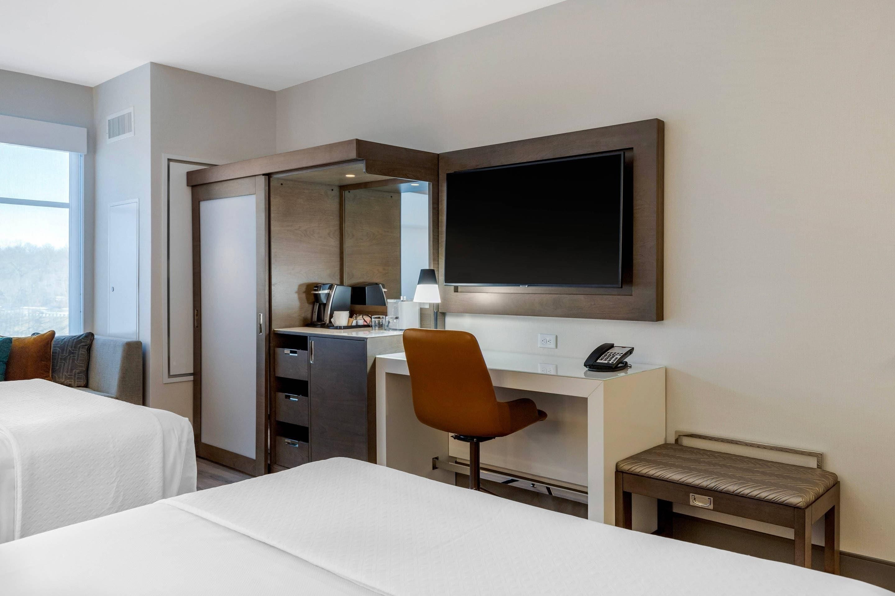 A hotel room with two beds, a desk, a chair, a small table, a mirror, and a TV.