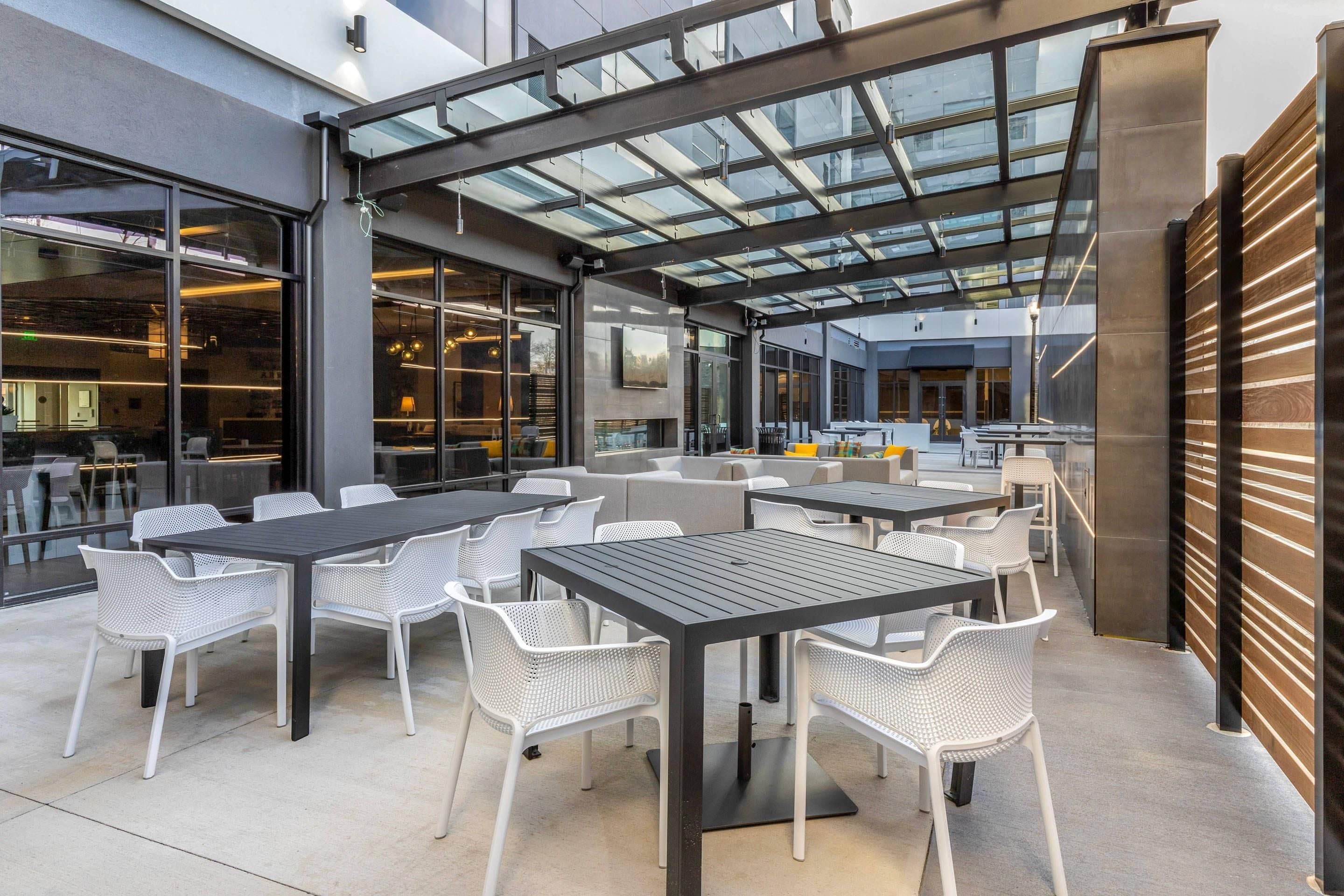 An outdoor seating area with tables and chairs, a glass ceiling, and a view of the interior.