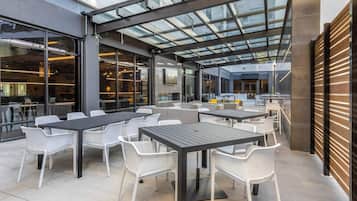An outdoor seating area with tables and chairs, a glass ceiling, and a view of the interior.