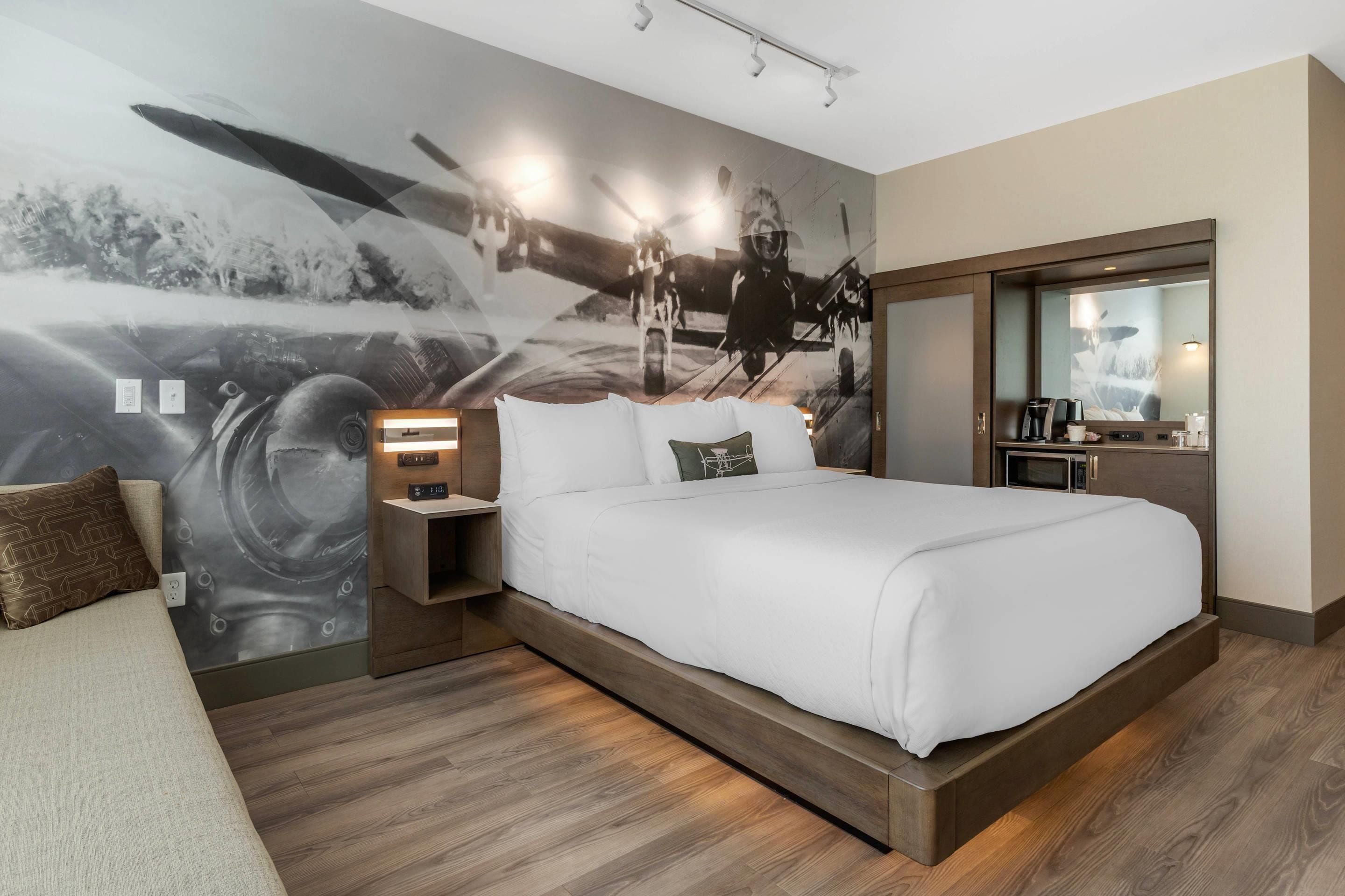 A modern hotel room with a large bed, a nightstand with a lamp, a built-in TV, and a large mirror.