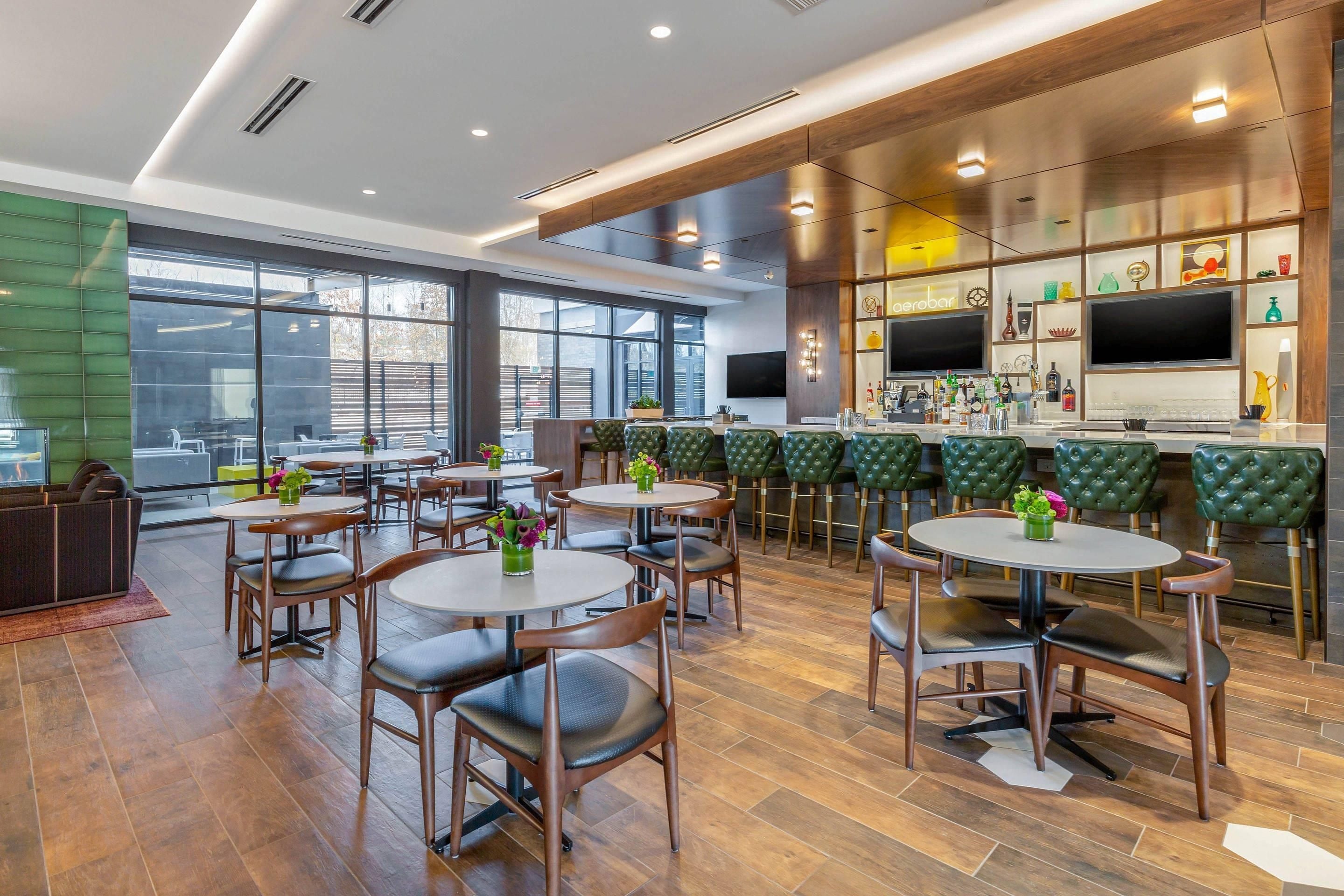 Un área de bar moderna con mesas redondas, sillas con cojines verdes y una variedad de botellas y vasos en las estanterías.
