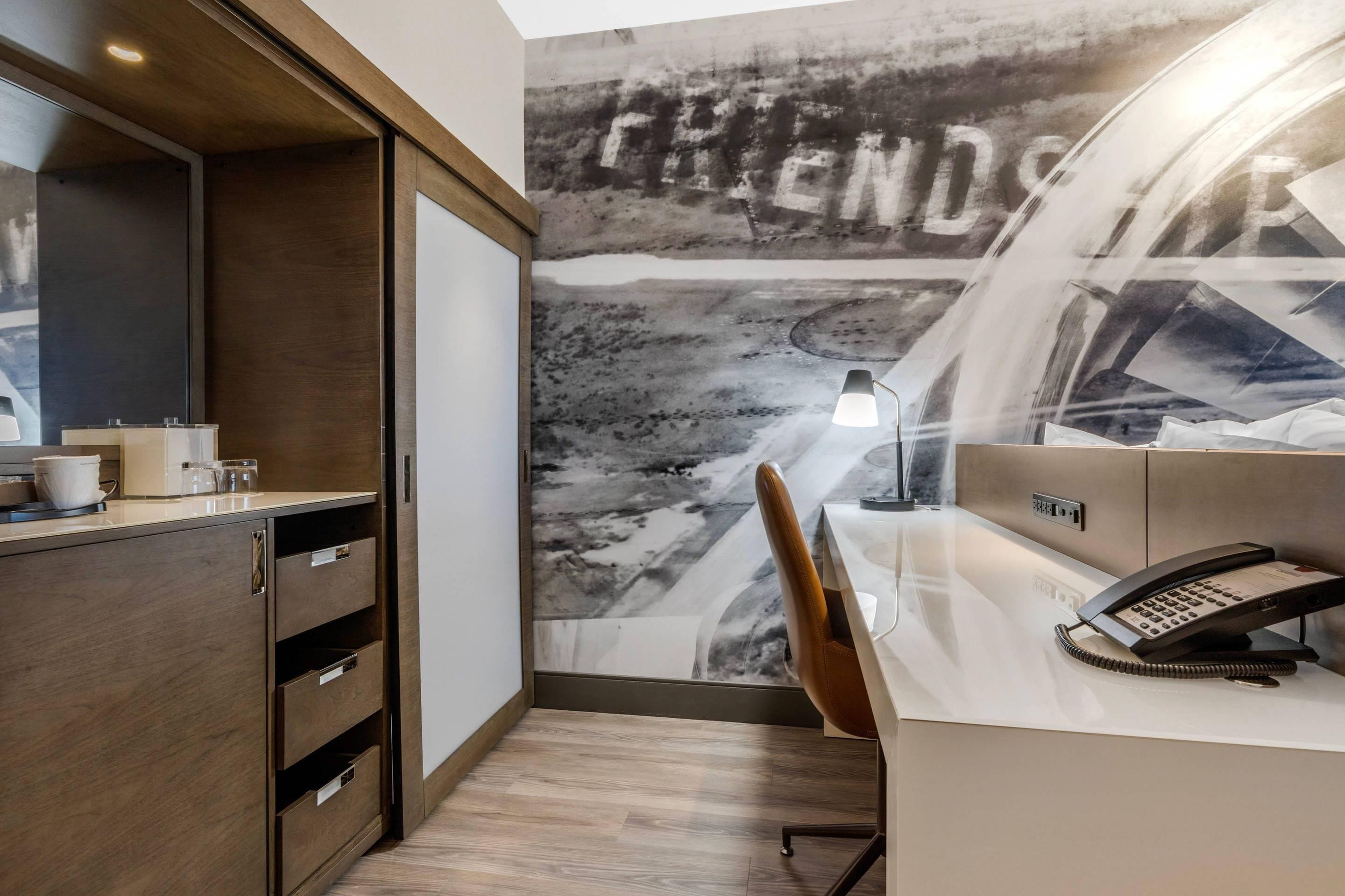 Habitación de hotel con escritorio, teléfono, cafetera y pared con un mural de 'FRIENDS'.