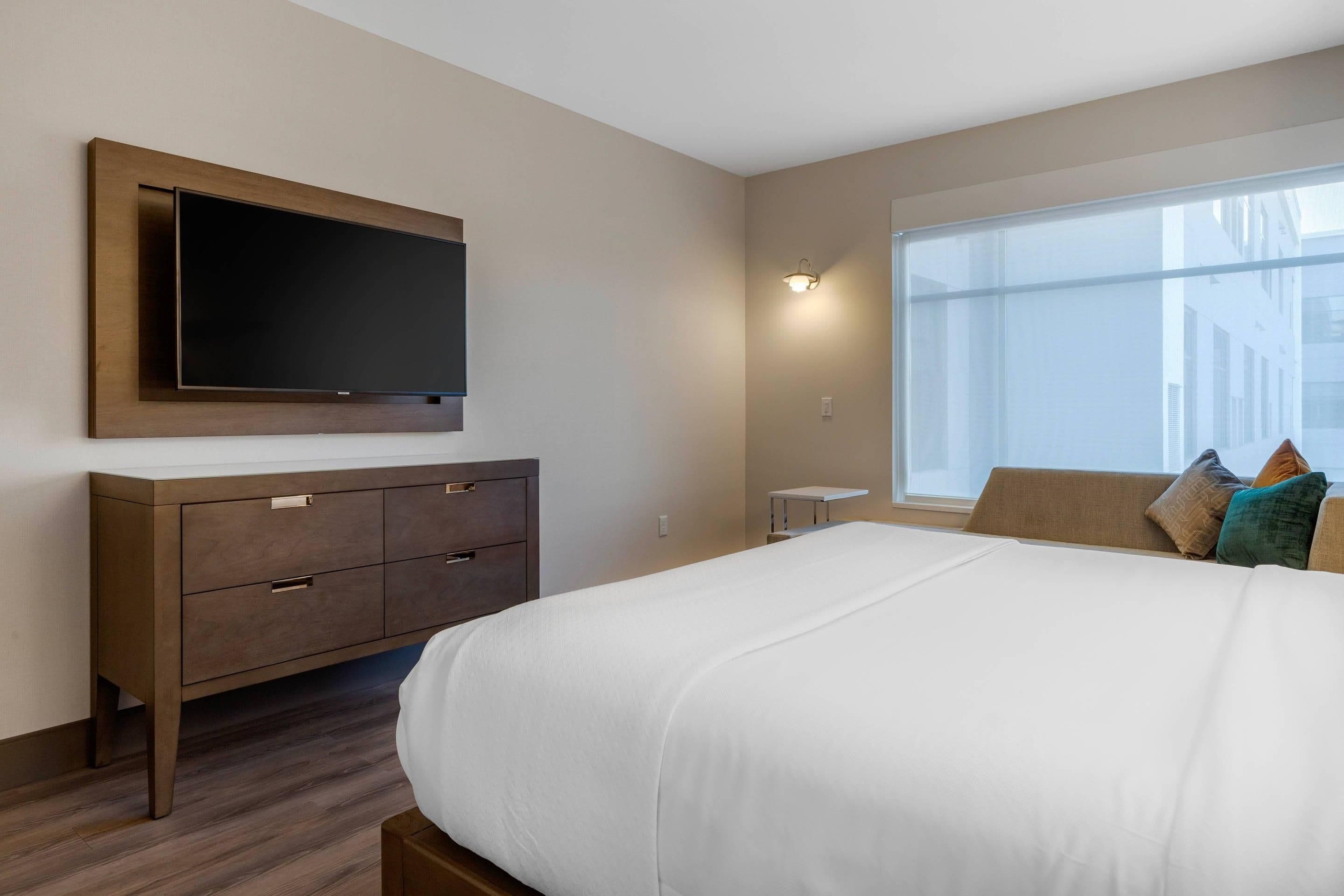 Habitación de hotel con una cama grande, un televisor de pantalla plana montado en la pared, una mesita de noche y una ventana con vistas a edificios.