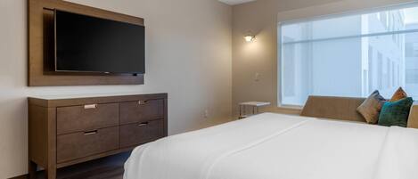 Habitación de hotel con una cama grande, un televisor de pantalla plana montado en la pared, una mesita de noche y una ventana con vistas a edificios.