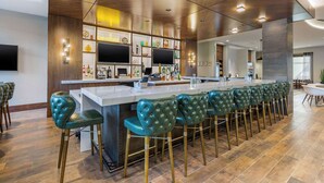 Un área de bar moderna con una barra larga, taburetes de bar tapizados en verde y un techo de madera con iluminación empotrada.