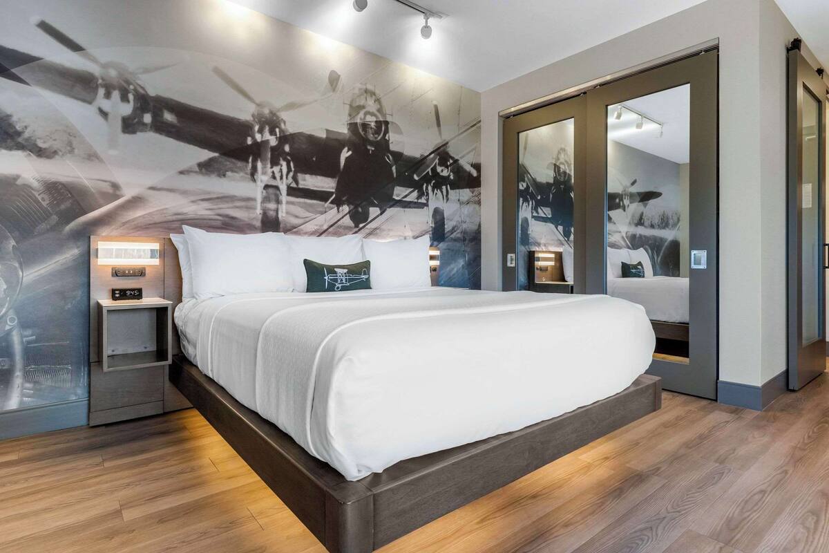 A modern bedroom with a large bed, a nightstand, a wall-mounted clock, and a mirror.