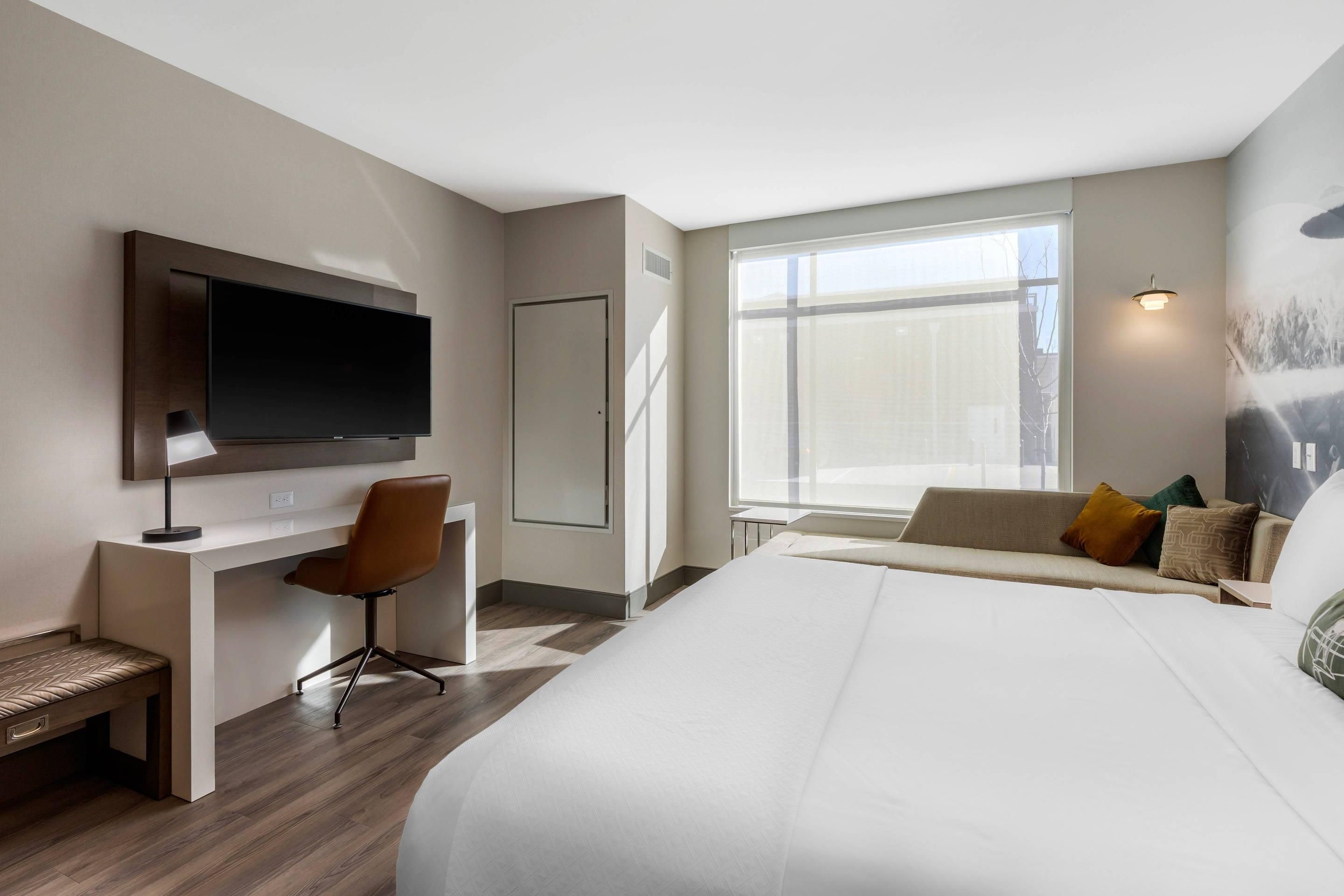 A modern hotel room with a large bed, a desk with a chair, a flat-screen TV, and a large window providing a view of the city.
