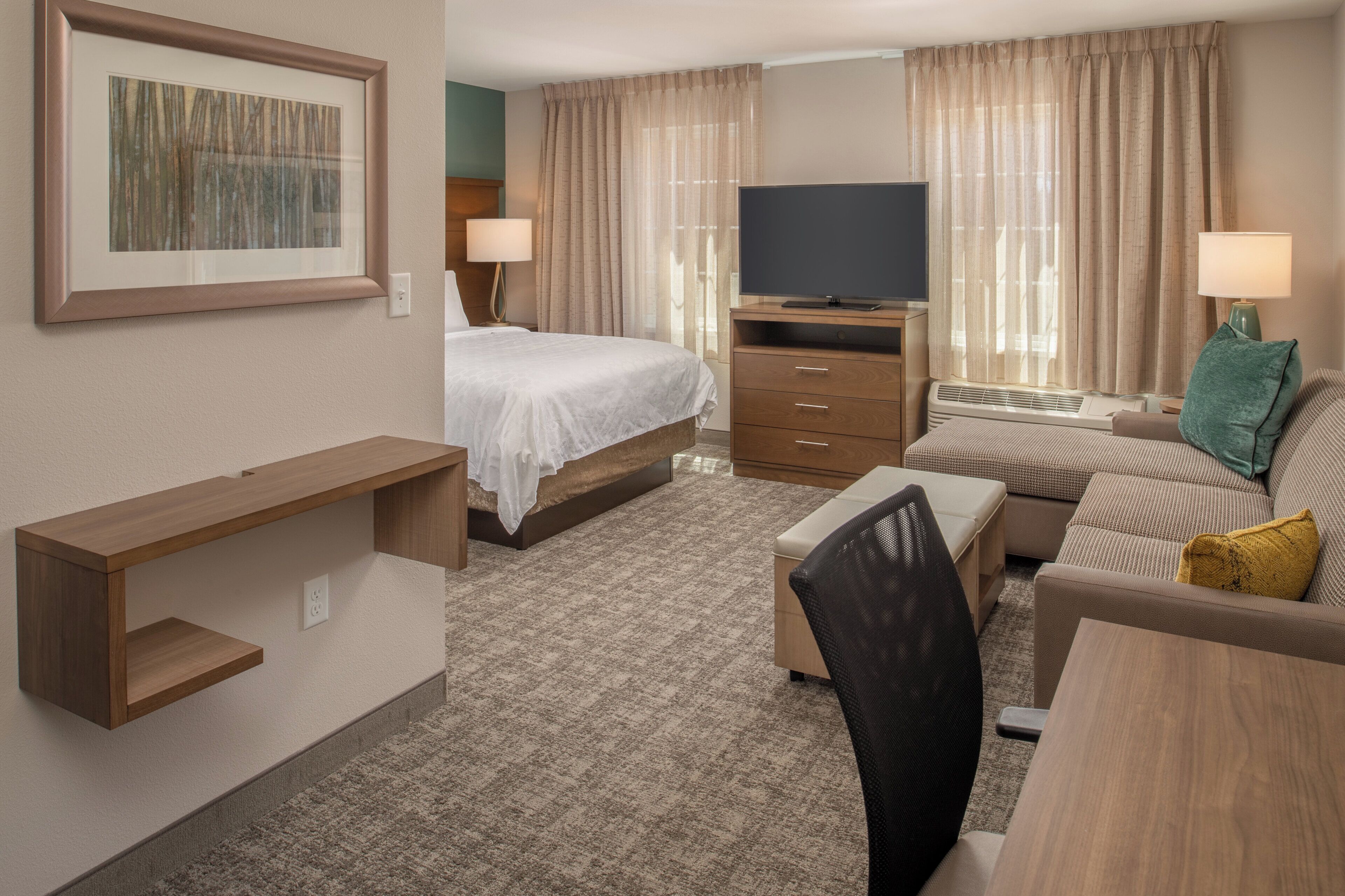 Habitación de hotel con cama, escritorio, silla, sofá, televisor y ventana con cortinas.