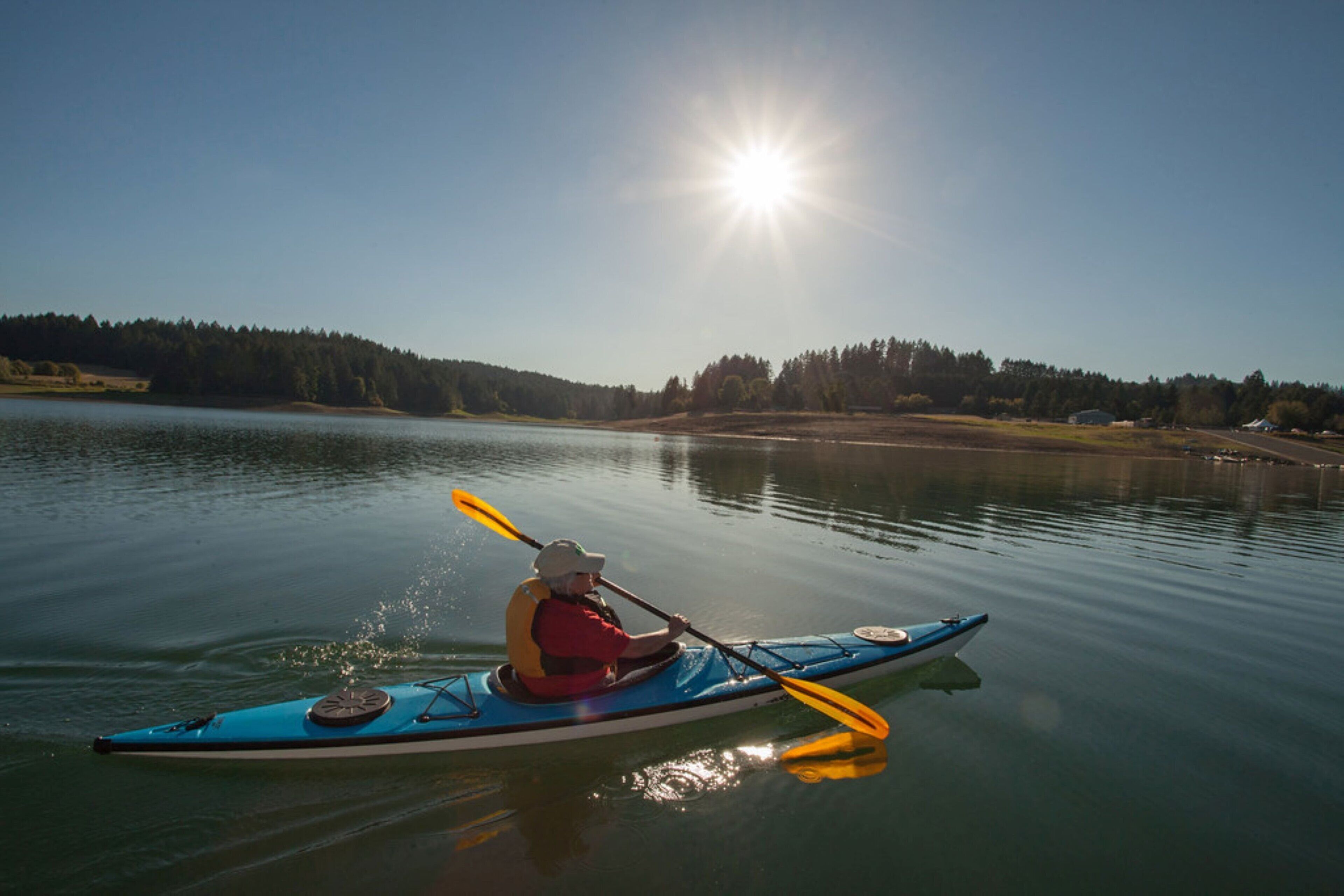 Un kayakista en kayak azul sobre aguas tranquilas, con cielo despejado y sol brillante.