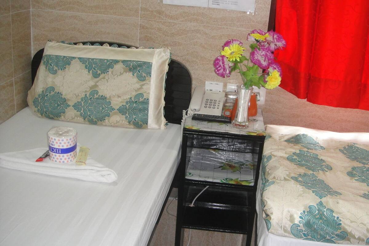 A hotel room with a bed, a bedside table with a telephone, a vase with flowers, and a tray with a cup and tissues.